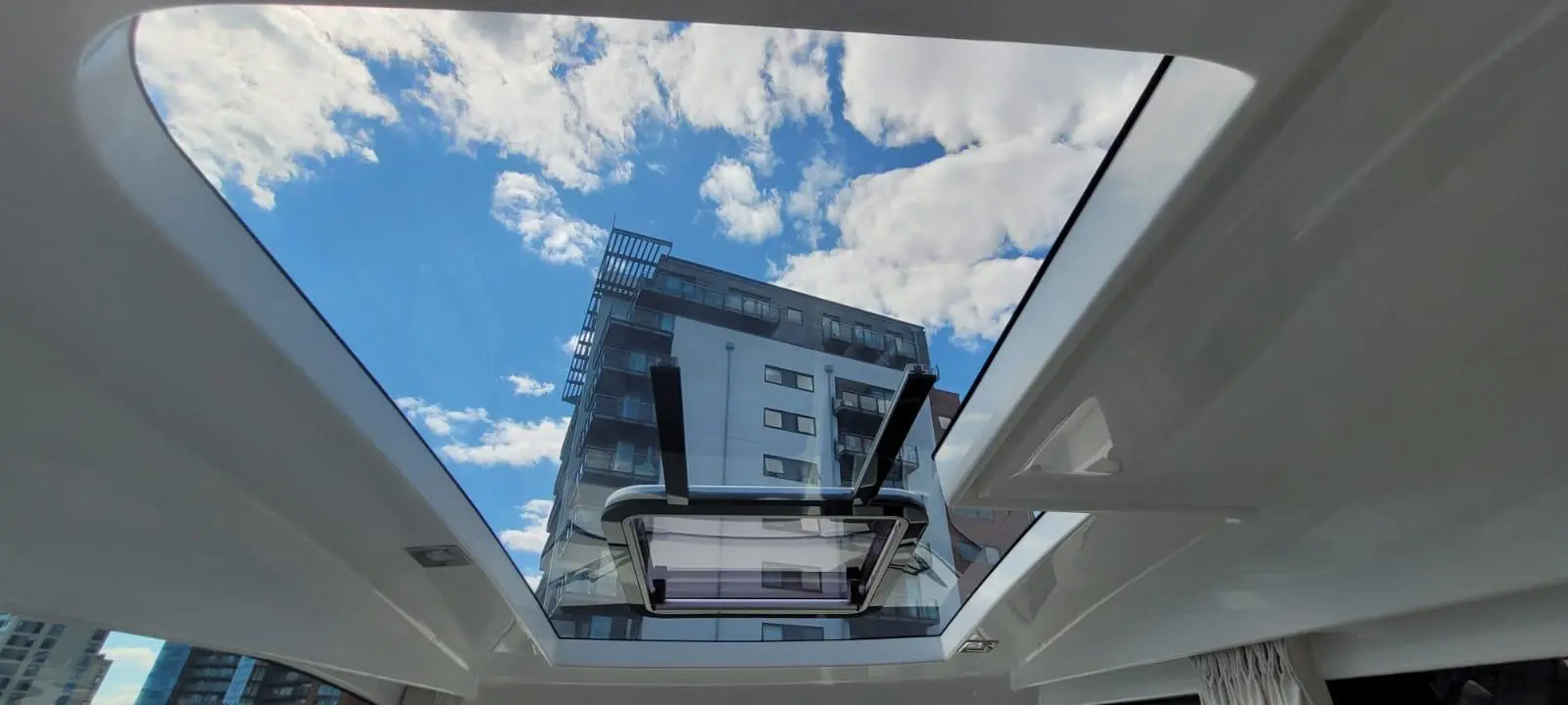 Sky view through a boat’s sunroof, featuring fluffy clouds and a modern building reflected in the glass.